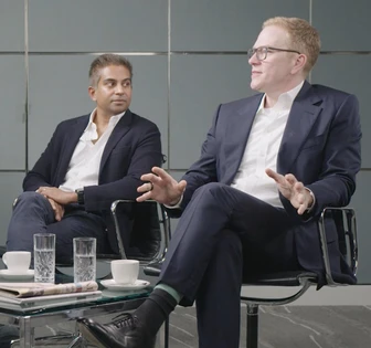 Two well-dressed men in business suits are engaged in conversation, seated at a table with glasses and a newspaper nearby, in what appears to be a professional setting