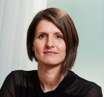 The image shows Adinah Shackleton, a middle-aged woman with shoulder-length brown hair, wearing a black jacket, and looking directly at the camera with a serious expression