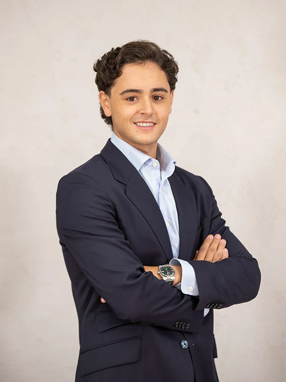 The image shows a young, well-dressed man wearing a navy blue suit and a light blue shirt, standing with his arms crossed and a warm smile on his face. The filename indicates his name is Juan Bedouka De La Chica