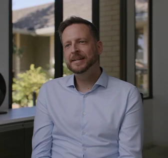 The image shows a smiling man with brown hair and a beard, wearing a light blue button-up shirt, standing in front of a window with a scenic view outside