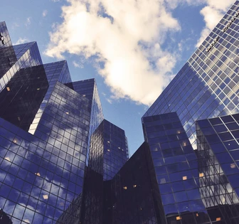 The image shows a striking urban skyline with tall, modern glass skyscrapers reflecting the cloudy blue sky above