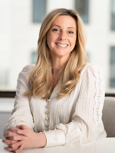 The image shows Alicia Daly, a smiling woman with long blonde hair wearing a white lace-trimmed blouse, posing in a professional setting