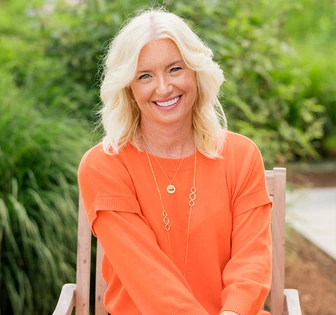 The image shows a smiling middle-aged woman with long, blonde hair wearing a bright orange sweater and necklace, seated in a garden setting