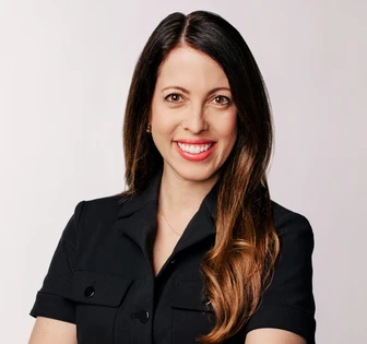 The image shows a headshot of Raina, a woman with long dark hair and a warm smile, wearing a black blazer against a plain background
