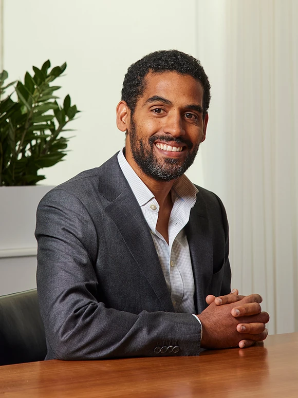 The image shows Michael Pote, a smiling middle-aged African American man wearing a gray suit and a checkered shirt, sitting at a desk in a professional setting