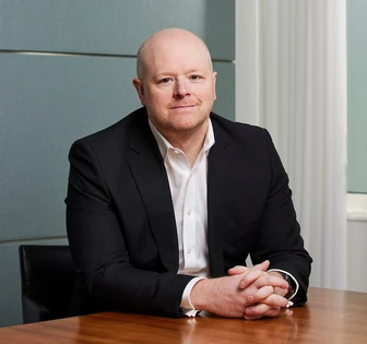 The image shows Ian Jackson, a middle-aged man, seated at a desk and looking directly at the camera. He is wearing a black suit and white shirt, with an expression of seriousness on his face