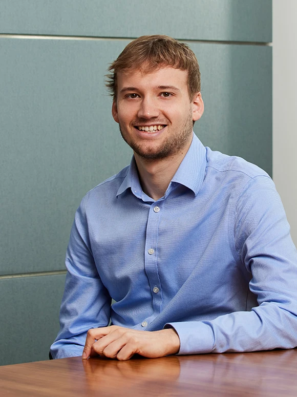 The image shows a smiling young man in a blue dress shirt sitting at a desk, with a plain grey background