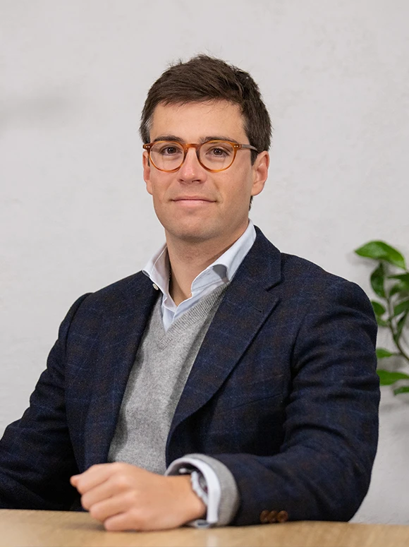 The image shows Gustavo Martinez, a professional-looking man wearing glasses and a suit, sitting at a desk and looking directly at the camera