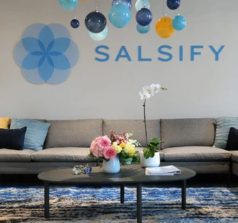 A bright, modern office space with hanging decorative balloons, a Salsify logo, a couch, a coffee table, and a vase of colorful flowers