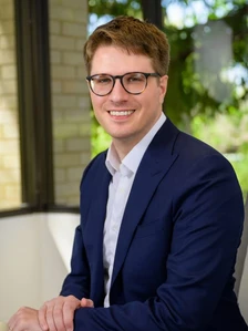 A smiling man in a navy blue suit and glasses, standing in front of a brick building and greenery, with the filename indicating his name is Sergio Rinaudo