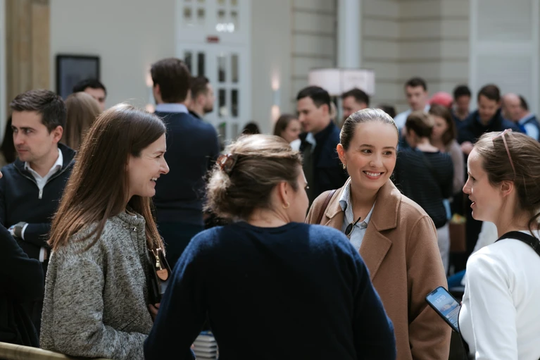A group of smartly dressed people, some engaged in conversation, stand in an indoor setting, suggesting a professional or social event