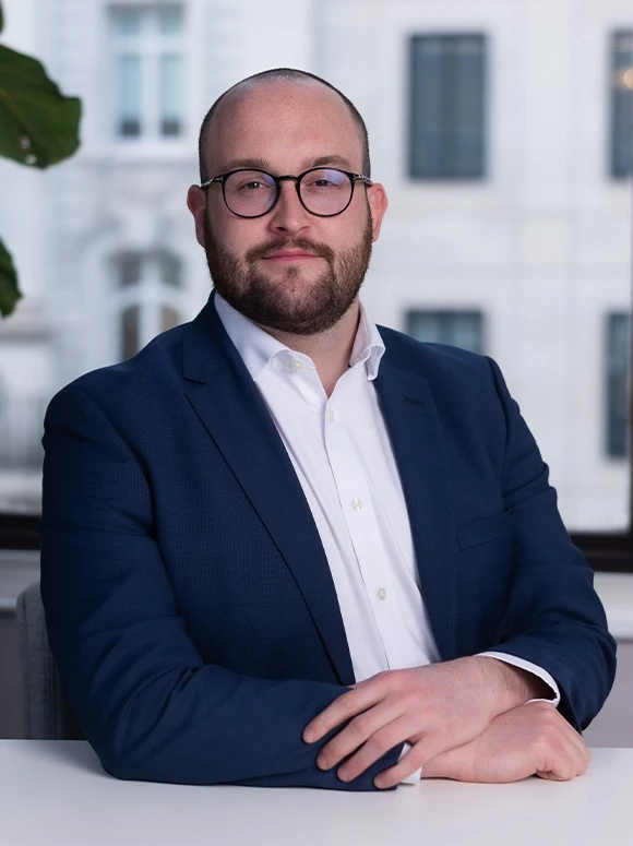 A smiling, middle-aged man with a beard, wearing glasses and a navy blue suit, sitting at a desk in a professional setting
