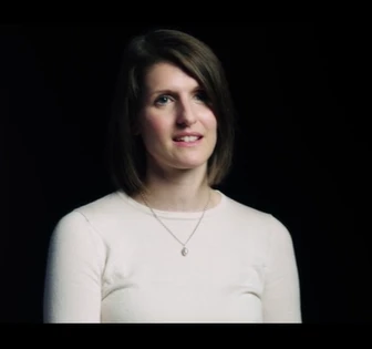 The image shows the portrait of a thoughtful-looking woman with dark hair wearing a white top against a dark background