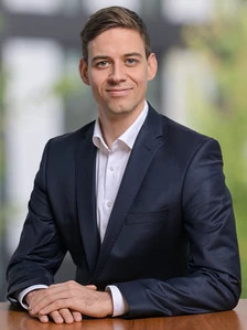 The image shows Timon Muler Gastell, a smartly dressed professional man in a suit, posing for a headshot against a blurred natural background