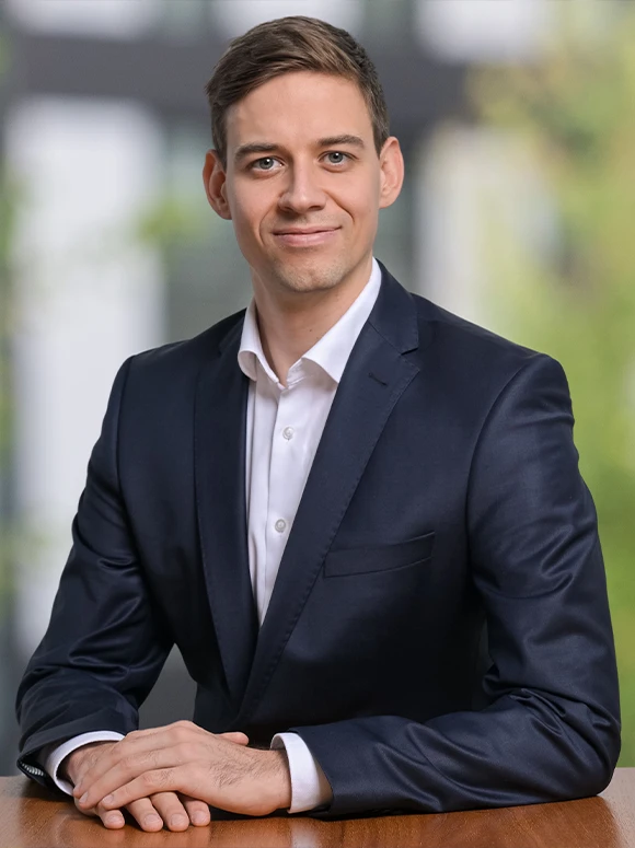 The image shows Timon Muler Gastell, a smartly dressed professional man in a suit, posing for a headshot against a blurred natural background
