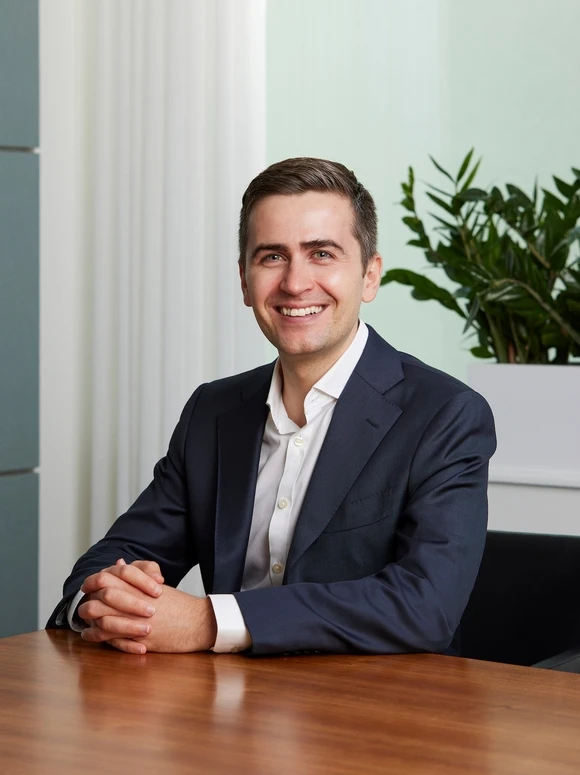 The image shows a smiling professional man named Stefan Ghinescu, wearing a dark suit and sitting at a desk in a room with plants in the background