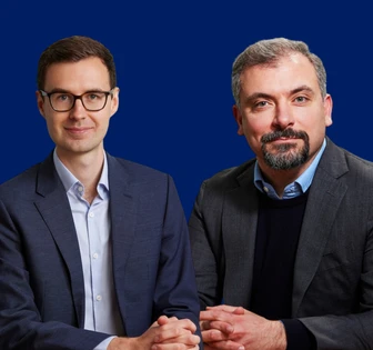 Two men, BR and RB, stand together in formal business attire against a blue background, appearing to have a friendly, professional interaction