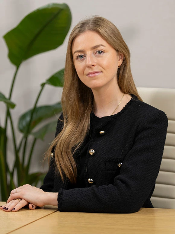 A professional-looking woman with long brown hair wearing a black jacket, sitting at a desk with a large green leaf in the background