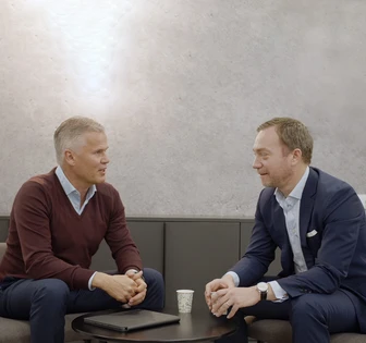 Two middle-aged men in formal attire, one in a burgundy sweater and the other in a suit, engaged in a conversation at a table