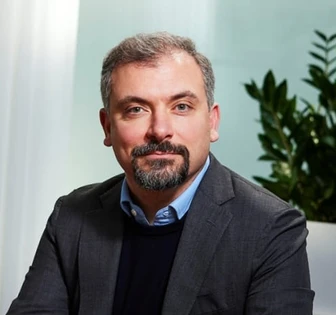 A bearded man with a serious expression, wearing a suit and sitting in front of a green background, identified as Riccardo Basile