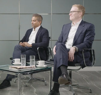 Two men in business attire sitting in chairs at a table with cups and glasses, likely in a professional setting