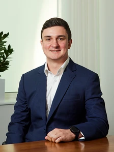 Jonny Tovey, a young professional in a navy blue suit, sitting at a desk and smiling