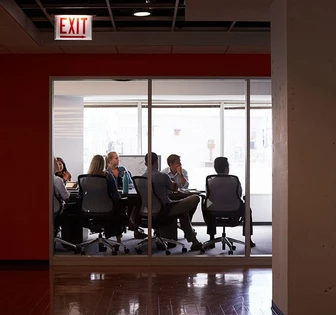 The image shows a group of professionals in business attire, seated around a conference table in a modern office setting with an illuminated "EXIT" sign overhead