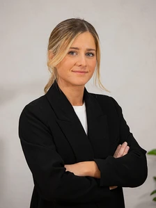 The image shows a professional-looking woman with blonde hair wearing a black blazer, standing with her arms crossed in a neutral pose