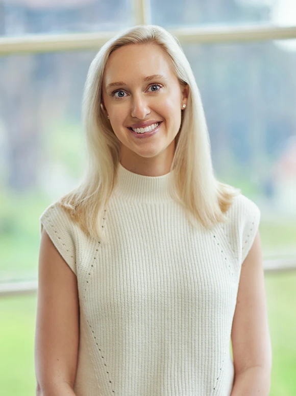 A smiling middle-aged woman with long blonde hair wearing a white sweater, against a blurred natural background