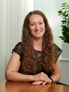 The image shows Orlanda Graham, a woman with long, wavy brown hair, smiling warmly while seated at a desk in what appears to be an office or home setting