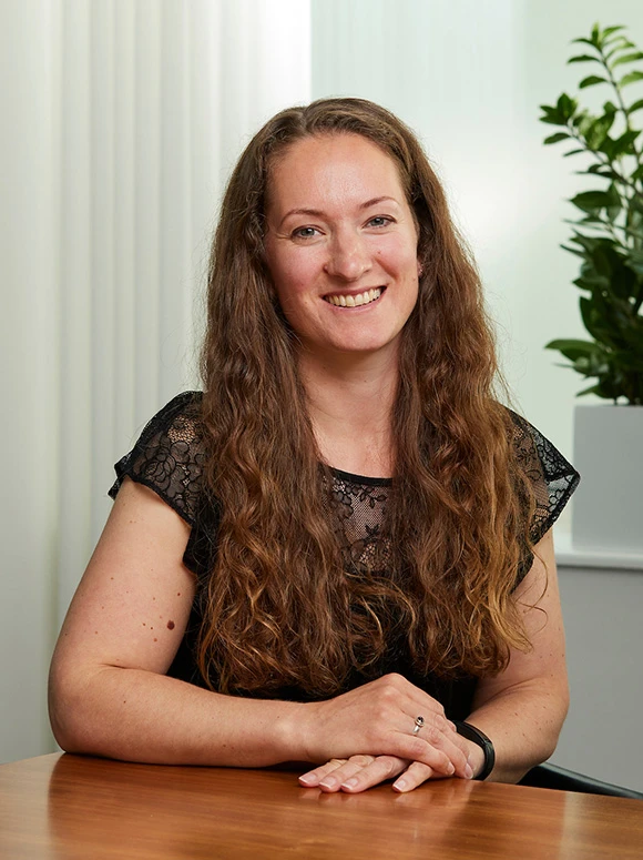 The image shows Orlanda Graham, a woman with long, wavy brown hair, smiling warmly while seated at a desk in what appears to be an office or home setting