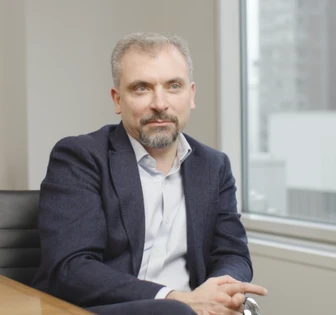 The image shows a well-dressed man with gray hair and a beard, sitting at a desk and gazing thoughtfully out a window