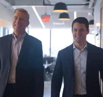 Two well-dressed, smiling businessmen walk together in a modern office setting, suggesting a partnership or collaboration