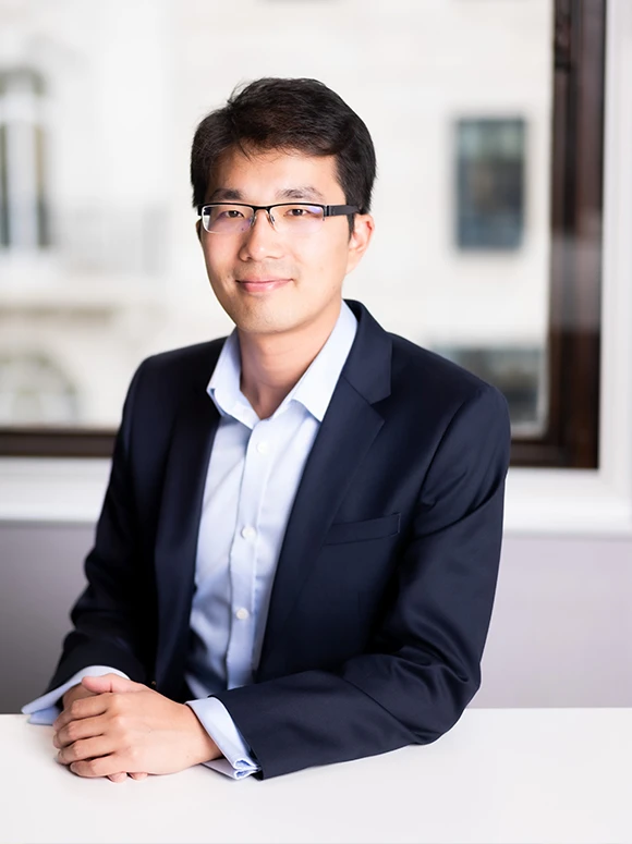 The image shows a smiling man in a suit and glasses, sitting at a desk and gazing directly at the camera