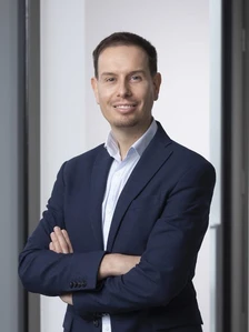 The image shows Eddy Perrier, a smiling man in his 30s or 40s, wearing a navy blue suit and a light-colored shirt, standing with his arms crossed in a professional pose