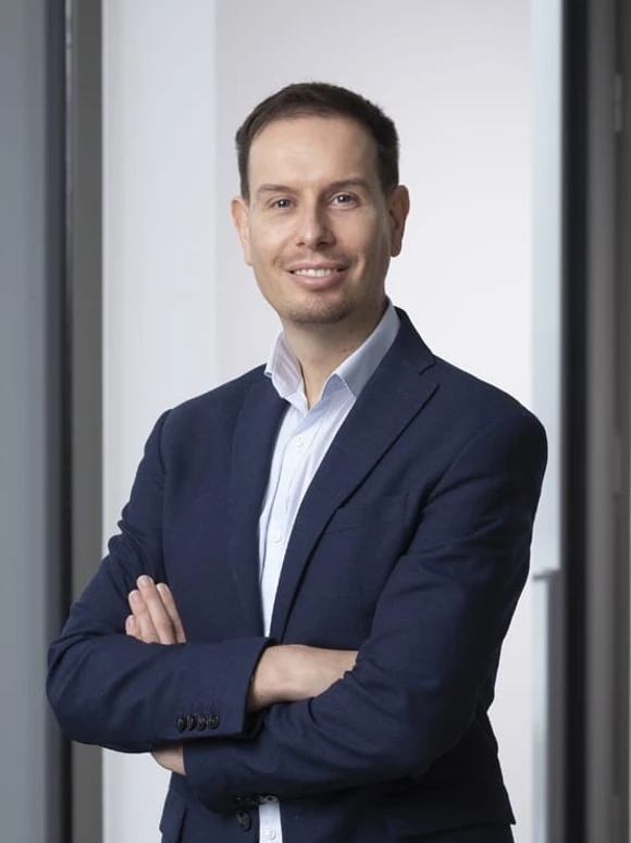 The image shows Eddy Perrier, a smiling man in his 30s or 40s, wearing a navy blue suit and a light-colored shirt, standing with his arms crossed in a professional pose
