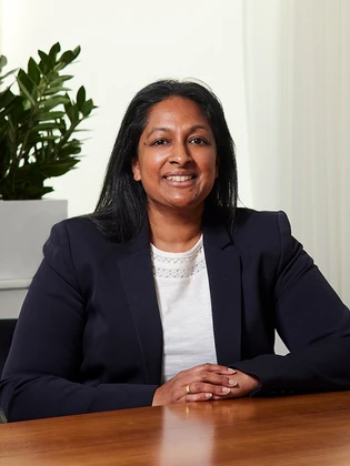 Ajaya Nair, a smiling professional woman in a black suit, sitting at a desk against a white background