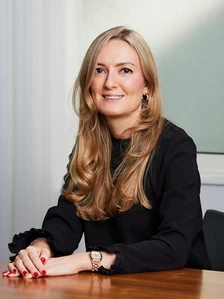 The image shows a smiling woman with long, wavy blonde hair wearing a black sweater, seated at a desk and looking directly at the camera