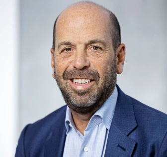 A portrait of Jeff Berkowitz, a middle-aged man with a beard, wearing a blue suit and checkered shirt, smiling warmly at the camera