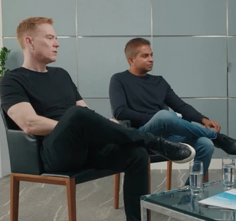 Two men seated in chairs having a conversation in an office setting, with one wearing a black top and the other in a blue top