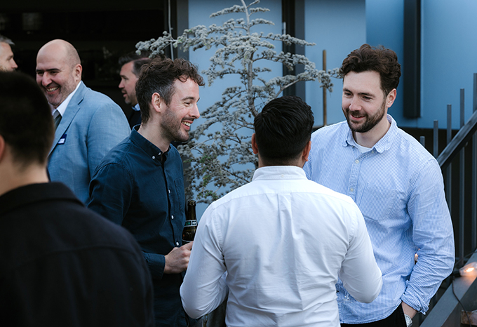 The image shows a group of five young men chatting and interacting in a snowy outdoor setting, with a decorated evergreen tree in the background