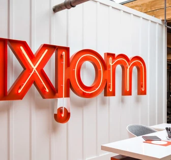 The image shows a large red neon sign displaying the word "Axiom" mounted on a white wooden wall, with a modern office desk and workspace visible in the background