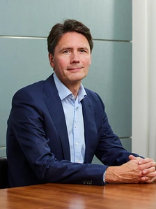 The image depicts a man in a navy blue suit, sitting at a desk and looking directly at the camera. He appears to be Peter Gibbs, based on the filename