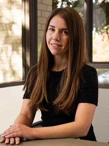 The image shows a smiling woman with long, brown hair sitting at a table and looking directly at the camera. The filename "Alexa Zahl Web (1)" suggests this is a portrait of Alexa Zahl