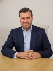 The image shows a middle-aged man in a navy blue suit sitting at a wooden desk. His name appears to be Jose Maria Palicio