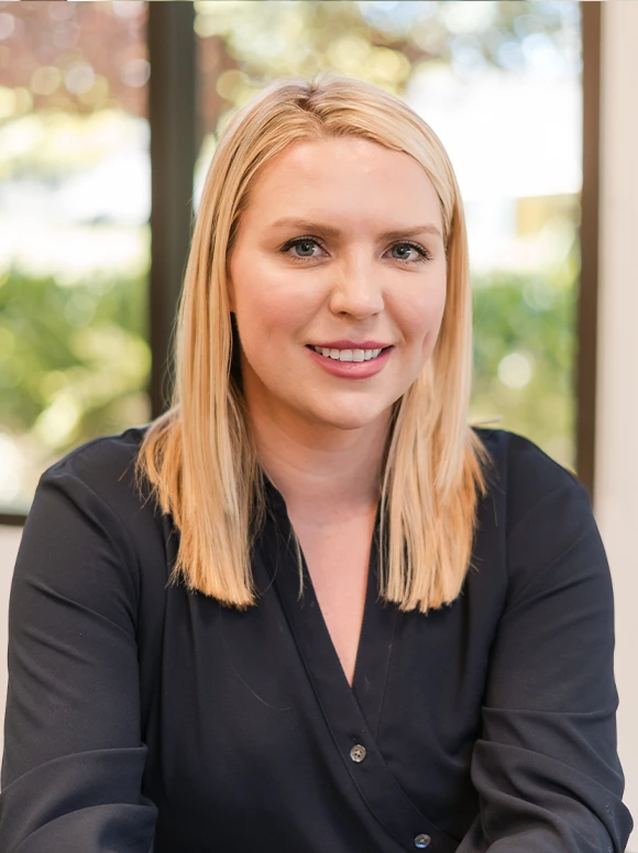 The image shows Megan Teixeira, a young woman with long blonde hair, wearing a black blazer, and smiling confidently in an outdoor setting