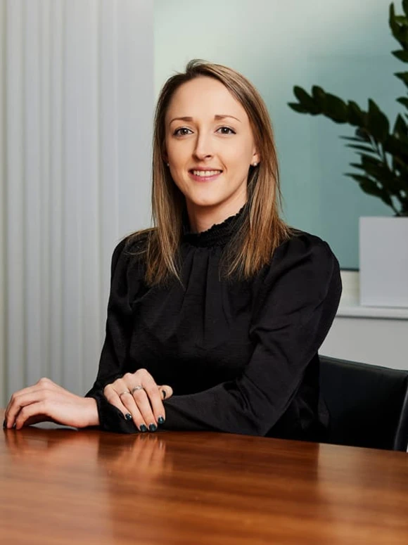 The image shows Lydia Galbraith, a professional-looking woman in her 30s or 40s, sitting at a desk in a formal office setting and smiling confidently at the camera