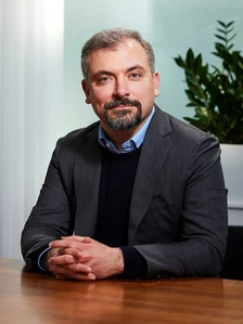 Riccardo Basile, a middle-aged man with a beard, sits at a desk in a professional office setting, dressed in a suit and tie