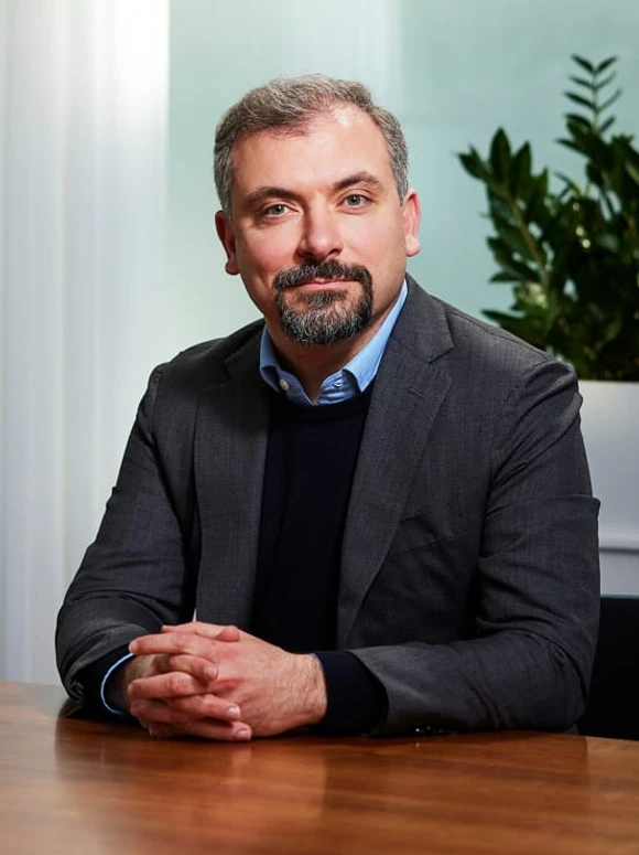 Riccardo Basile, a middle-aged man with a beard, sits at a desk in a professional office setting, dressed in a suit and tie