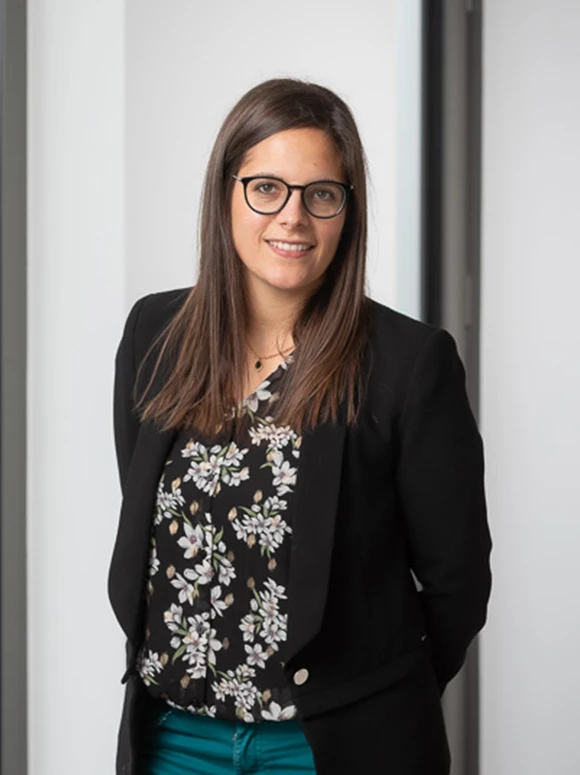 The image shows a smiling young woman with long brown hair wearing eyeglasses and a black blazer over a floral print blouse, standing in front of a light-colored wall